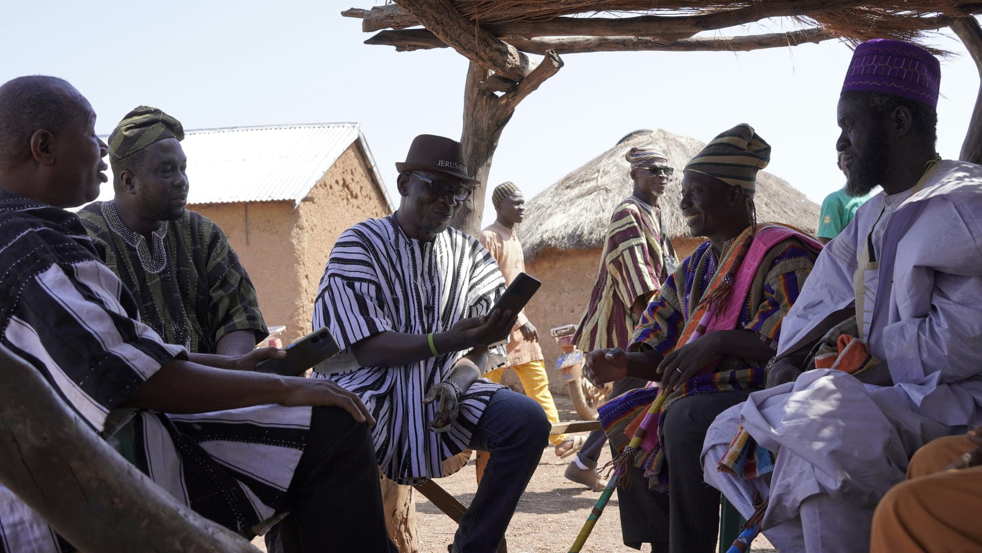 Village elders in traditional dress gathered in discussion, one holding a smartphone